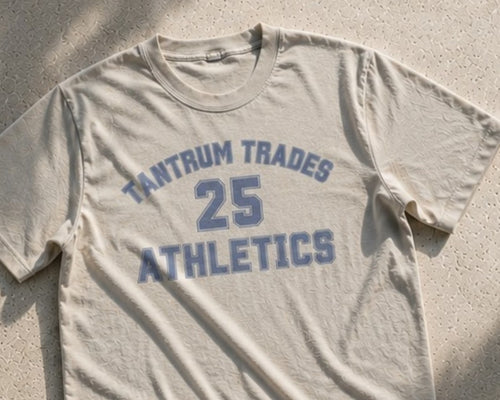 A steel grey crewneck t-shirt with 'TANTRUM TRADES 25 ATHLETICS' printed in a faded blue varsity font, laid flat on a textured concrete surface.