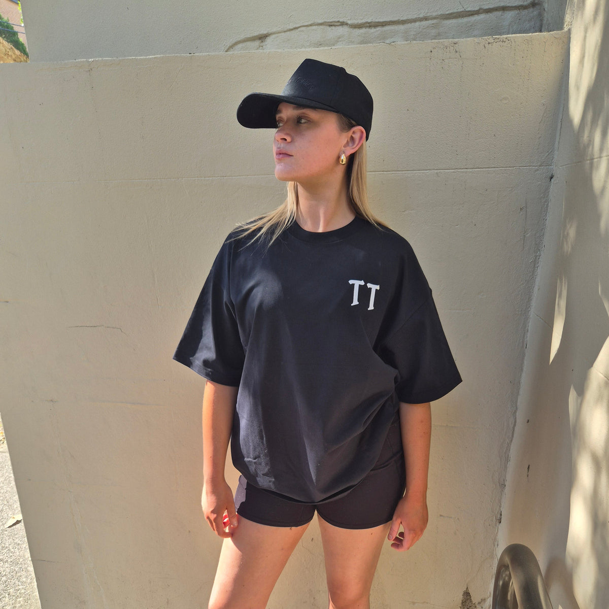 A woman wearing a black oversized t-shirt with a white 'TT' logo on the chest, black athletic shorts, a black baseball cap with black embroidered 'TANTRUM TRADES', white socks, and white sneakers, standing outdoors against a concrete wall.