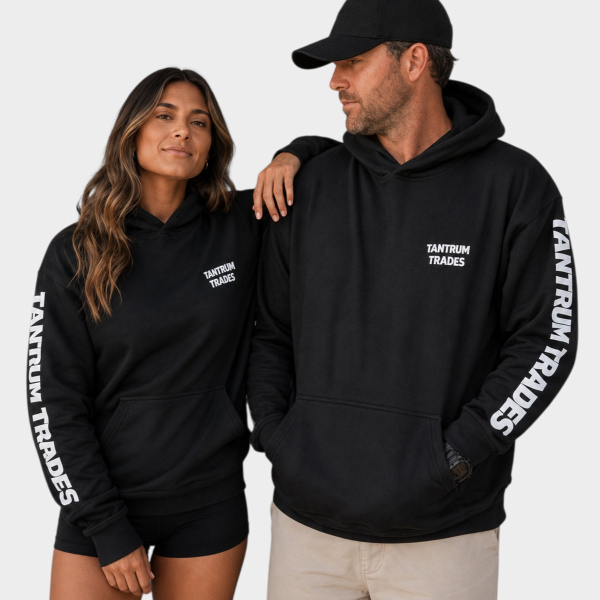 A man and a woman wearing matching black hoodies with white 'TANTRUM TRADES' text on the chest and down the sleeves, posed against a light gray studio background.