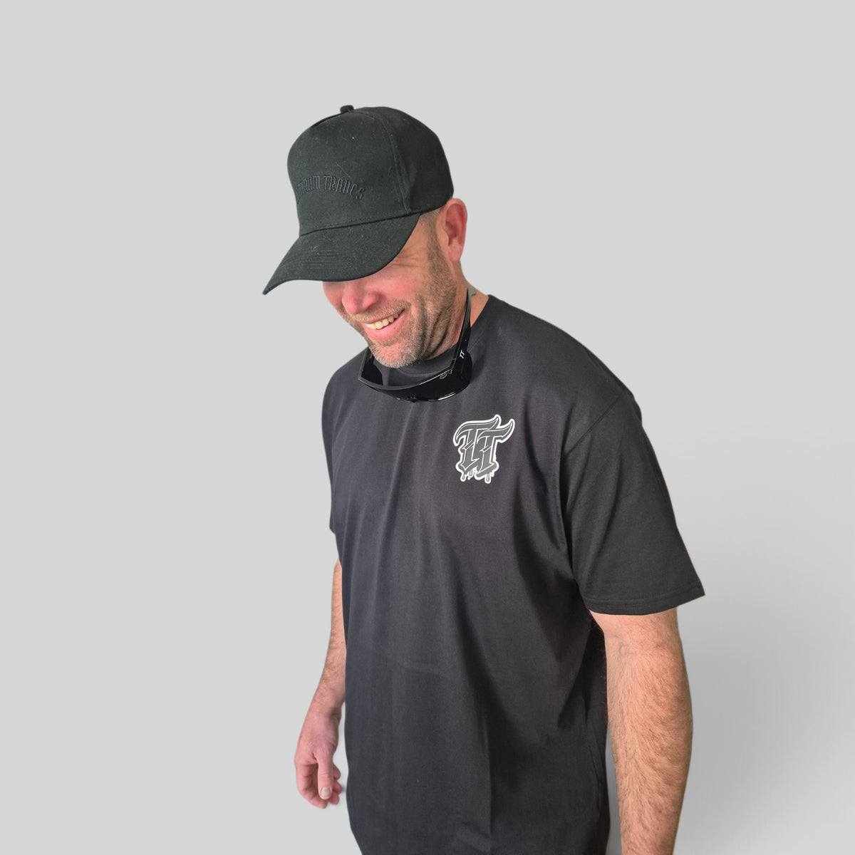 A man wearing a black Tantrum Trades baseball cap and a black t-shirt with a white and black graphic 'TT' logo on the left chest, photographed against a solid light gray background.
