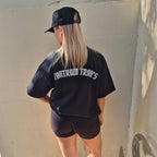 A woman viewed from behind wearing a black oversized t-shirt with 'TANTRUM TRADES' logo printed in white text on the back, paired with black shorts and a black 'Tantrum Trades' baseballhat, standing against a concrete wall.
