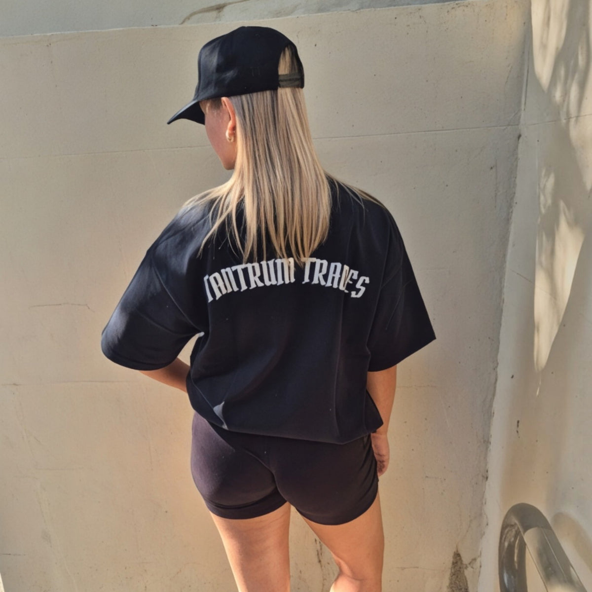 A woman viewed from behind wearing a black oversized t-shirt with 'TANTRUM TRADES' logo printed in white text on the back, paired with black shorts and a black 'Tantrum Trades' baseballhat, standing against a concrete wall.