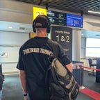 A man seen from behind wearing a black Tantrum Trade t-shirt with 'TANTRUM TRADES' printed in beige on the back, a black baseball cap, and carrying a black and gray backpack in an airport terminal.