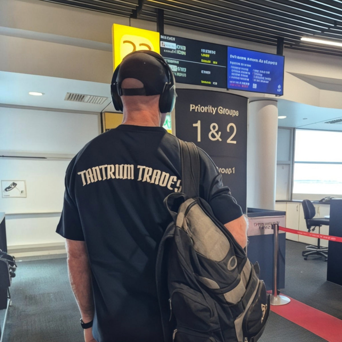 A man seen from behind wearing a black Tantrum Trade t-shirt with 'TANTRUM TRADES' printed in beige on the back, a black baseball cap, and carrying a black and gray backpack in an airport terminal.
