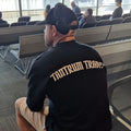A FIFO mine worker sitting in an airport waiting area wearing a black T-shirt with 'TANTRUM TRADES' logo printed across the back in a white font, along with a black Tantrum Trades cap and over-ear headphones.
