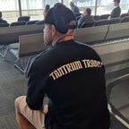 A FIFO mine worker sitting in an airport waiting area wearing a black T-shirt with 'TANTRUM TRADES' logo printed across the back in a white font, along with a black Tantrum Trades cap and over-ear headphones.