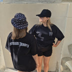 Two women standing outdoors against a concrete wall, both wearing black t-shirts. The woman on the left is seen from the back wearing a 'TANTRUM TRADES' logo bucket hat and a black 'TT' logo t-shirt with 'TANTRUM TRADES' printed in white.