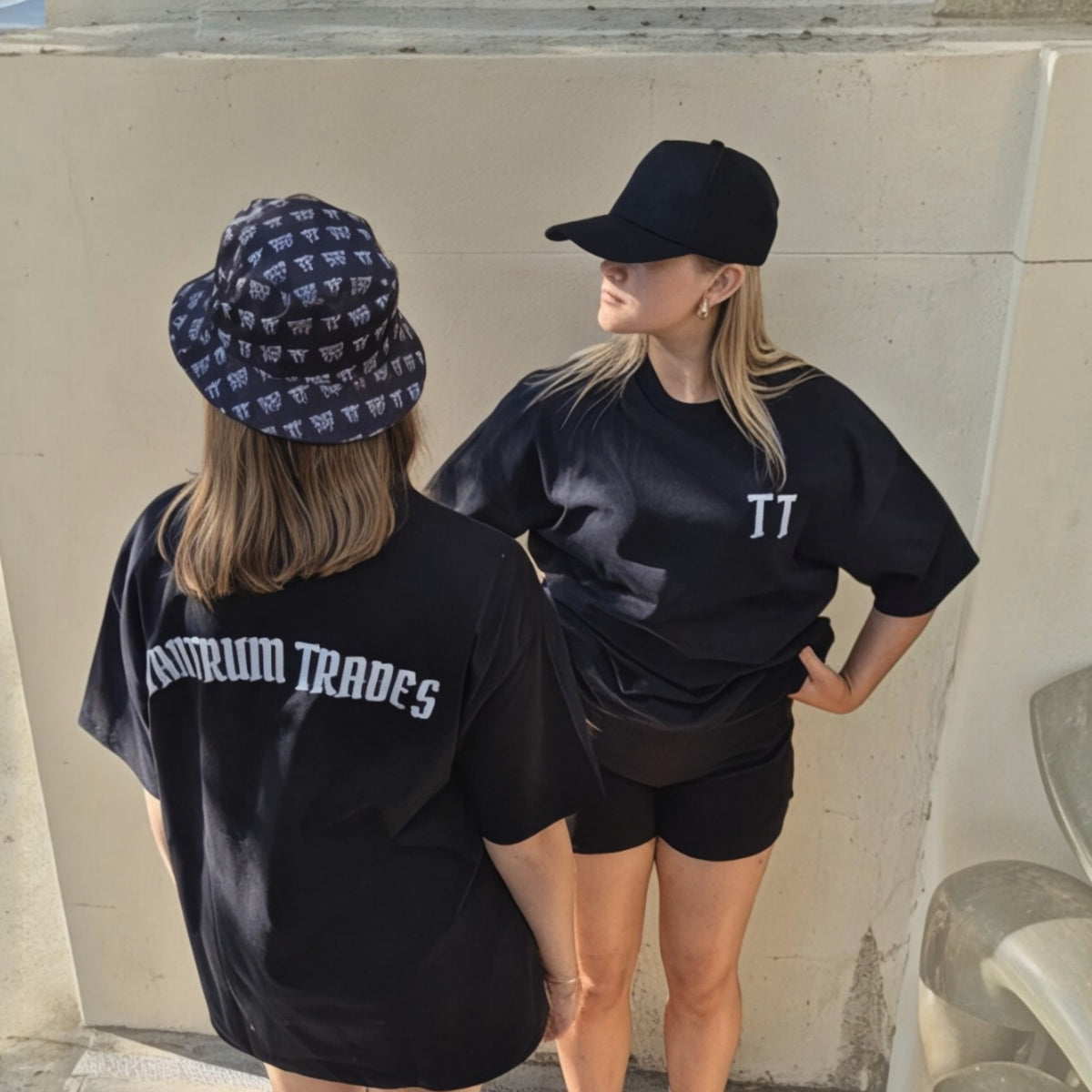 Two women standing outdoors against a concrete wall, both wearing black t-shirts. The woman on the left is seen from the back wearing a 'TANTRUM TRADES' logo bucket hat and a black 'TT' logo t-shirt with 'TANTRUM TRADES' printed in white.