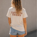 A woman viewed from behind wearing a cream-colored t-shirt with 'TANTRUM TRADES' printed in brown across the back, paired with light blue denim shorts, standing against a textured gray wall.