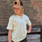 A woman wearing a cream-colored T-shirt with a small brown 'TANTRUM TRADES' logo on the left chest, a tan and black trucker hat, and dark brown shorts, standing in front of a rustic brick wall.