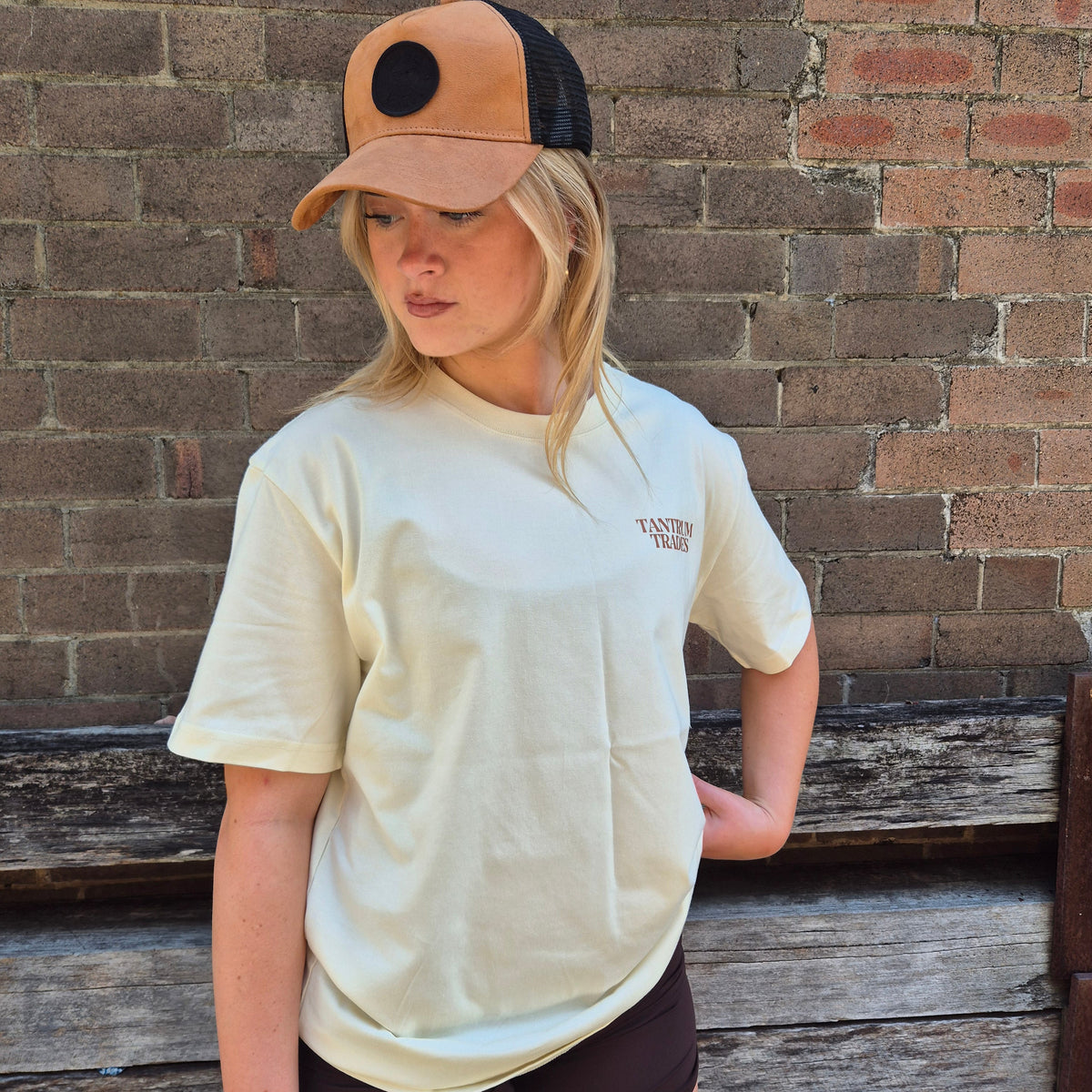 A woman wearing a cream-colored T-shirt with a small brown 'TANTRUM TRADES' logo on the left chest, a tan and black trucker hat, and dark brown shorts, standing in front of a rustic brick wall.