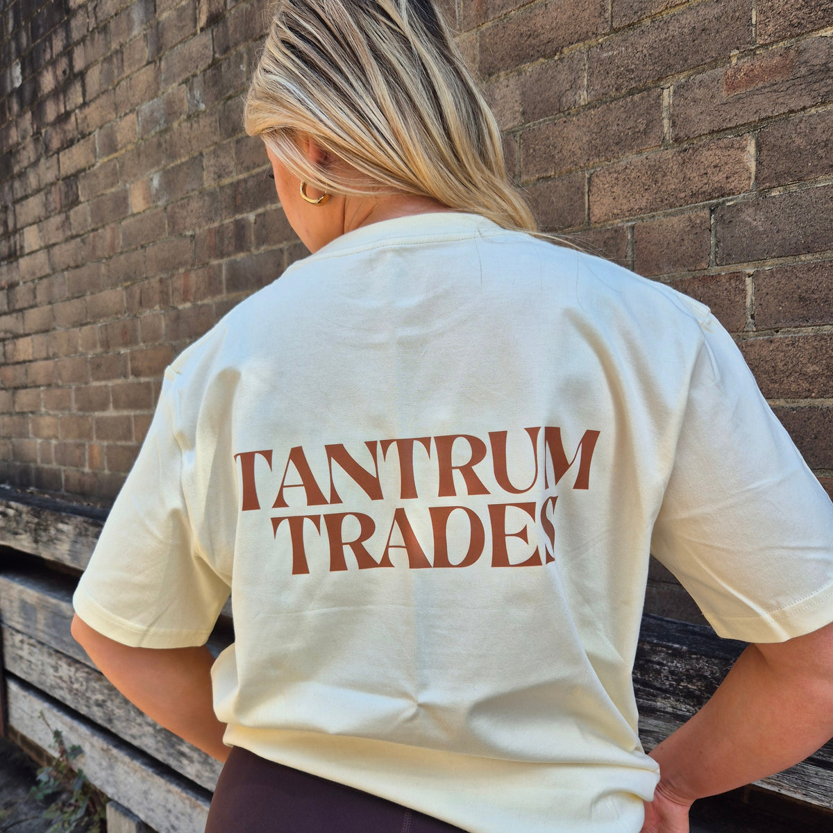 A woman viewed from behind wearing a cream-colored t-shirt with 'TANTRUM TRADES' printed in brown across the back, standing against a brick wall.