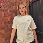 A woman with blonde hair stands against a red brick wall and a black metal gate, wearing a cream-colored oversized t-shirt with 'TANTRUM TRADES' printed in brown on the left chest, paired with brown shorts.