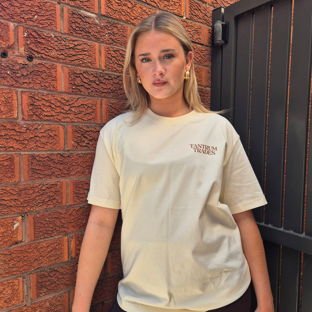 A woman with blonde hair stands against a red brick wall and a black metal gate, wearing a cream-colored oversized t-shirt with 'TANTRUM TRADES' printed in brown on the left chest, paired with brown shorts.