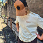 A person wearing a tan and black trucker hat and a cream t-shirt with 'TANTRUM TRADES' printed on the chest, standing outdoors against a brick wall.