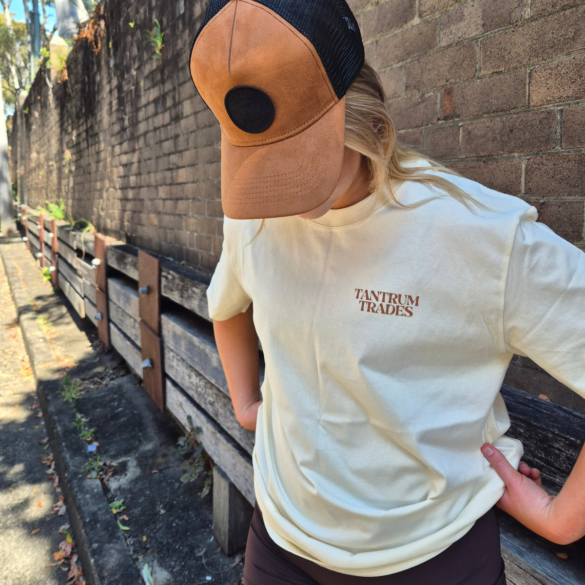 A person wearing a tan and black trucker hat and a cream t-shirt with 'TANTRUM TRADES' printed on the chest, standing outdoors against a brick wall.