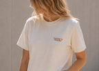 A woman wearing a cream-colored short-sleeve t-shirt with a small brown 'Tantrum Trades' logo on the left chest, paired with light-wash denim shorts, standing against a concrete wall.