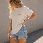 A woman wearing a cream-colored t-shirt with 'TANTRUM TRADES' logo printed in brown on the left chest, paired with light-wash denim shorts, standing against a textured gray wall.