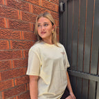 A woman wearing a cream-colored oversized 'TANTRUM TRADES' t-shirt and brown shorts, posing against a red brick wall and a dark metal gate.