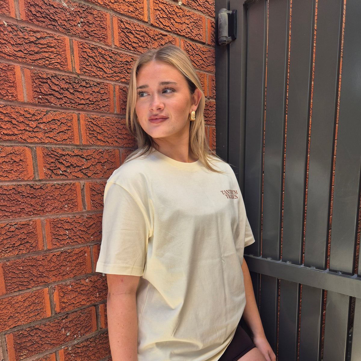 A woman wearing a cream-colored oversized 'TANTRUM TRADES' t-shirt and brown shorts, posing against a red brick wall and a dark metal gate.