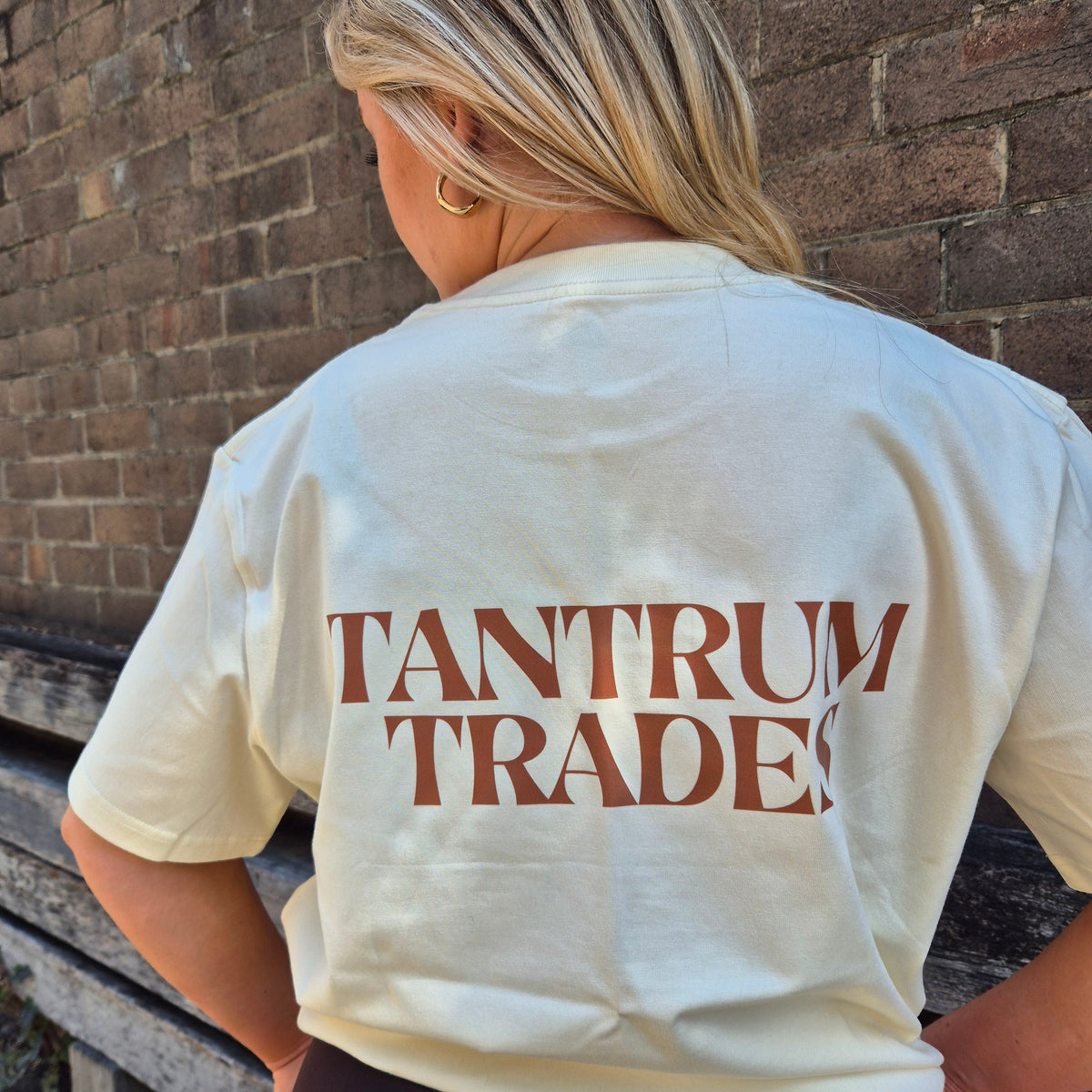 A woman viewed from behind wearing a cream-colored t-shirt with 'TANTRUM TRADES' printed in brown across the back, standing in front of a brick wall.