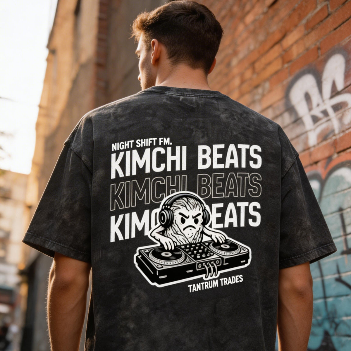 A man viewed from behind wearing an acid washed black graphic t-shirt with 'KIMCHI BEATS' and a DJ illustration printed in white on the back, standing in an outdoor alleyway.