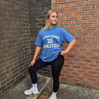 A woman posing outdoors in a blue athletic t-shirt with 'TANTRUM TRADES 25 ATHLETICS' printed in white, paired with black leggings and white sneakers, standing against a brick wall corner.