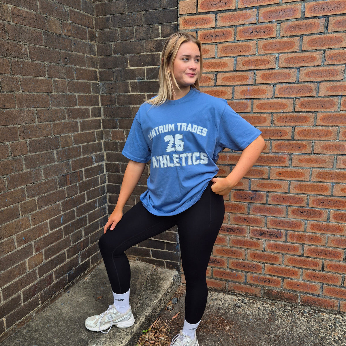 A woman posing outdoors in a blue athletic t-shirt with 'TANTRUM TRADES 25 ATHLETICS' printed in white, paired with black leggings and white sneakers, standing against a brick wall corner.