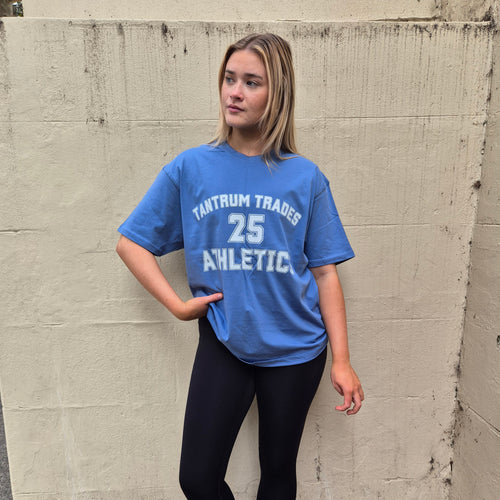 A woman stands against a concrete wall wearing a blue graphic t-shirt with 'TANTRUM TRADES 25 ATHLETIC' printed in white, paired with black leggings and white sneakers.