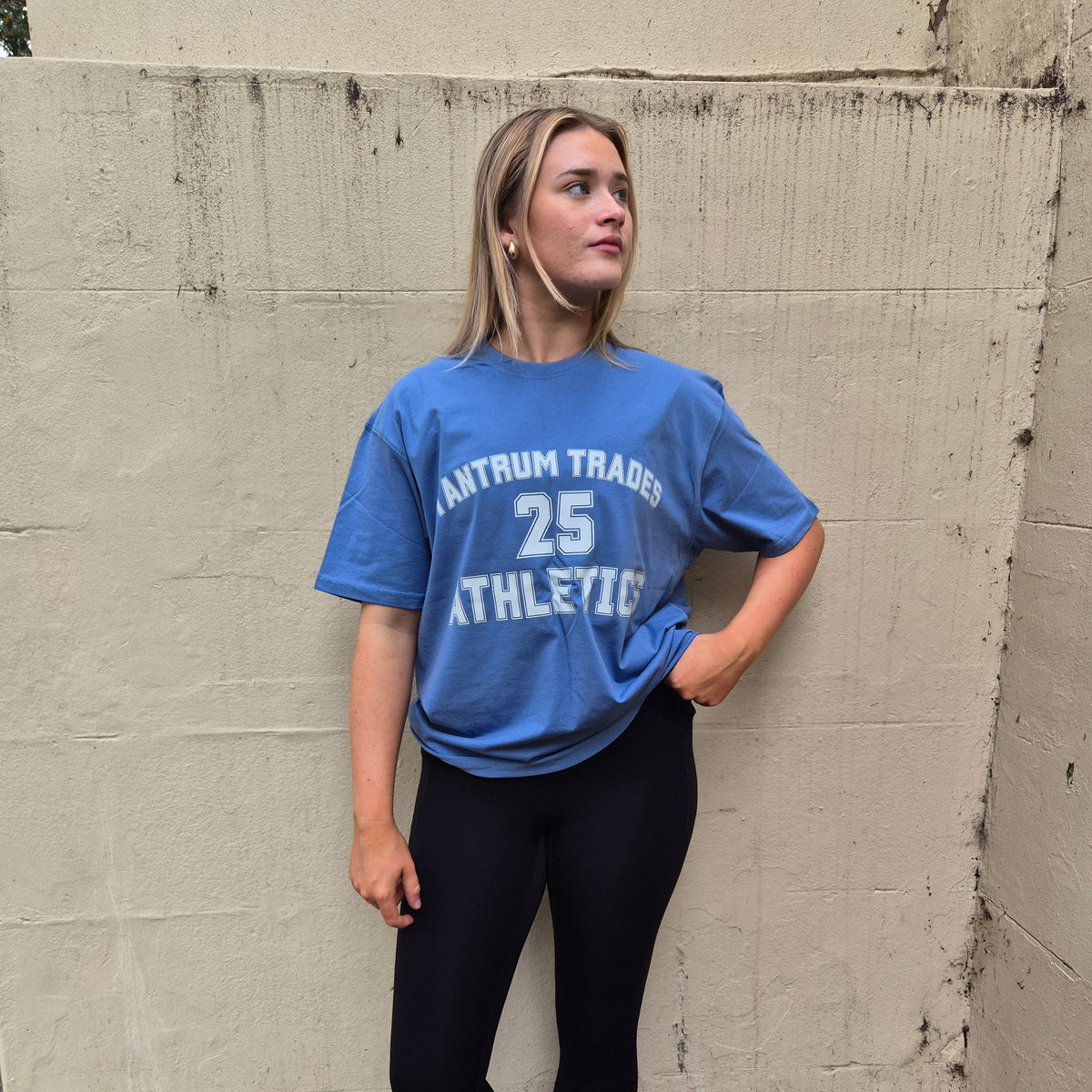 A woman stands against a concrete wall wearing a blue athletic t-shirt with 'TANTRUM TRADES 25 ATHLETIC' printed in white, paired with black leggings, white socks, and white sneakers.