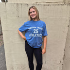 A young woman stands against a concrete wall wearing a blue athletic t-shirt with 'TANTRUM TRADE 25 ATHLETICS' printed in white, black leggings, white socks, and white sneakers.