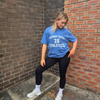 A woman posing in a blue athletic t-shirt with white lettering that reads 'TANTRUM TRADES 25 ATHLETICS', paired with black leggings, white socks, and white sneakers, standing in a brick alleyway.